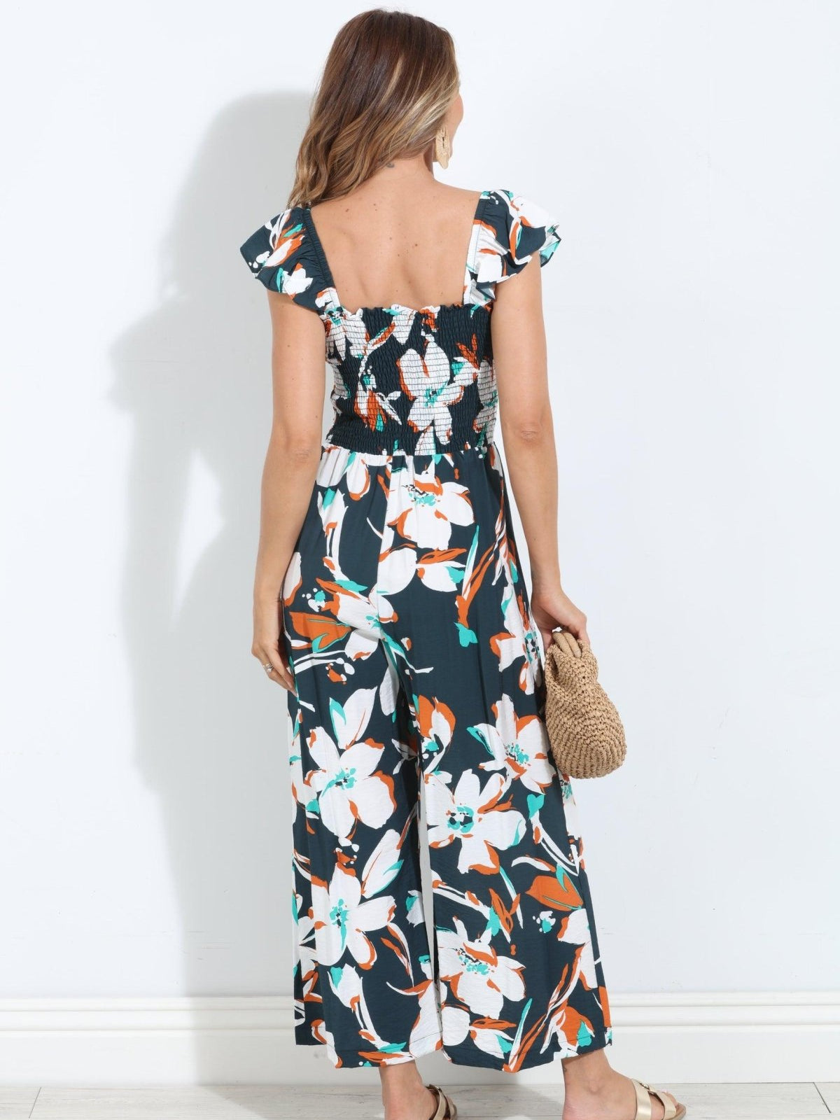backside of sleeveless floral jumpsuit on woman holding tan purse and in gold sandals