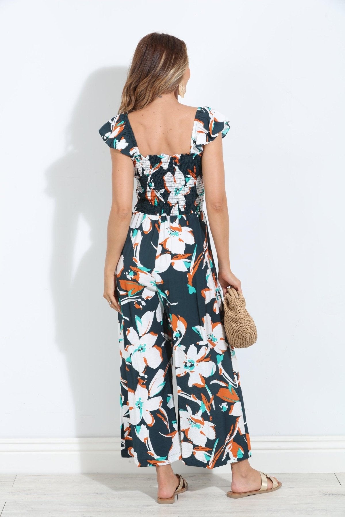 backside of sleeveless floral jumpsuit on woman holding tan purse and in gold sandals