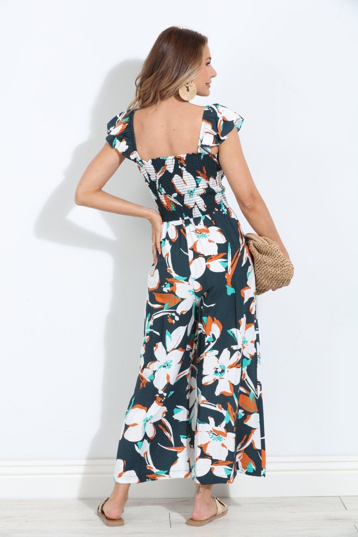 backside of sleeveless floral jumpsuit on woman holding tan purse and in gold sandals