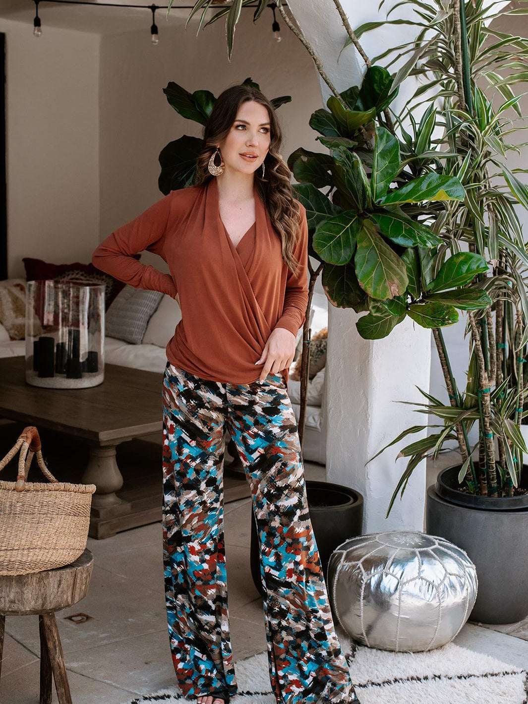 dark orange long sleeve blouse on woman in multicolored pants next to plants and home decor