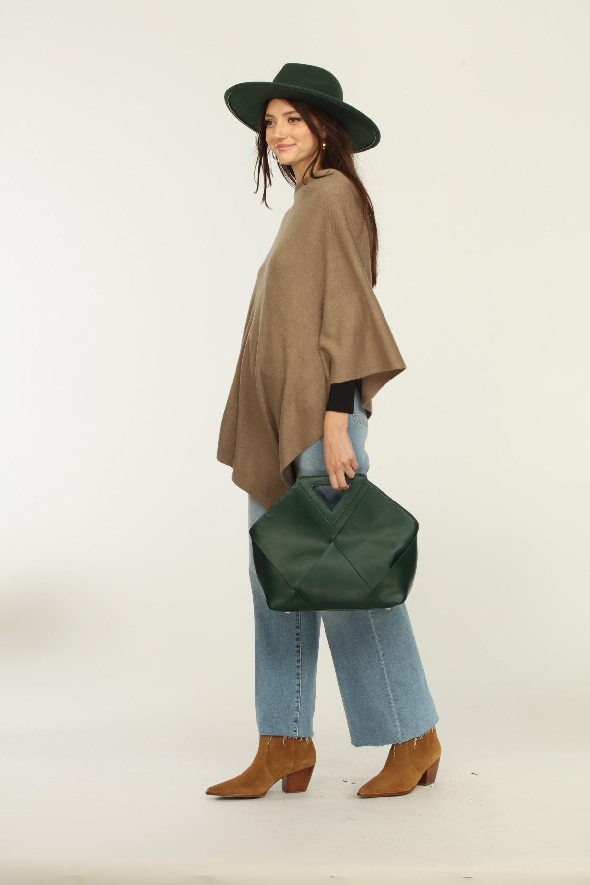 side view of woman wearing a hat, tan poncho, jeans, and tan boots holding green purse