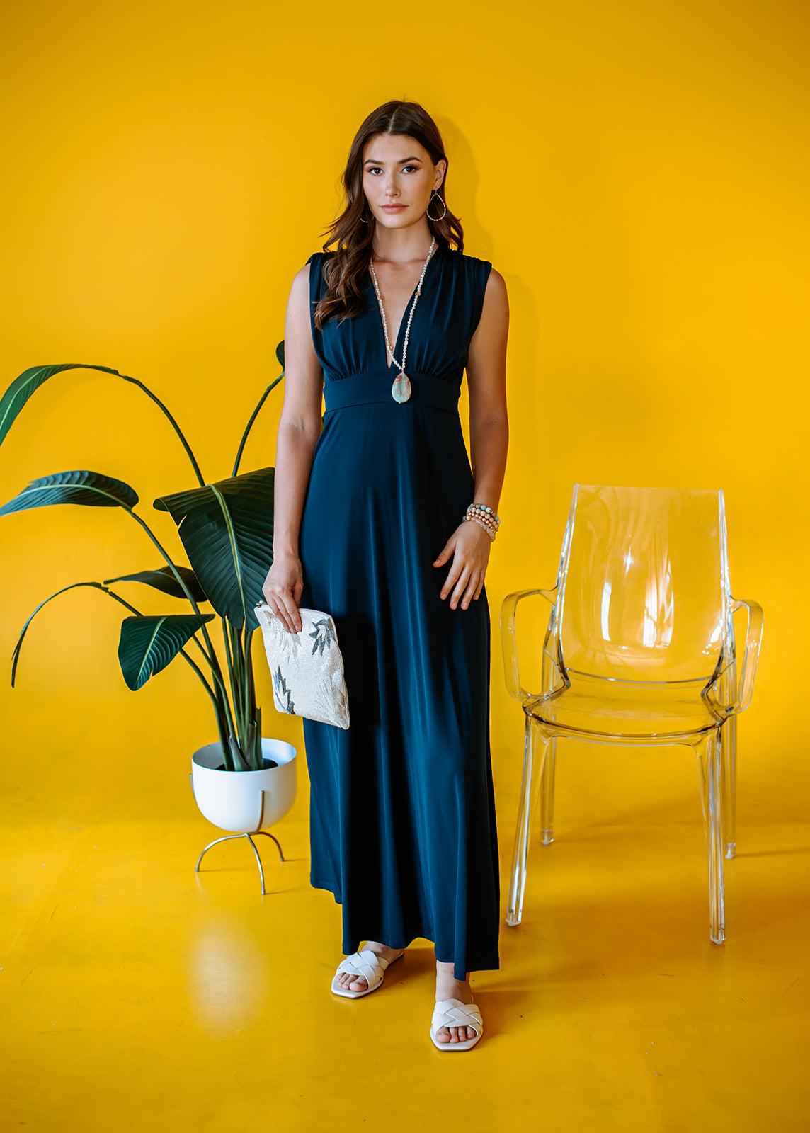 navy sleeveless maxi dress with deep neckline and side slit on woman holding a purse with yellow background