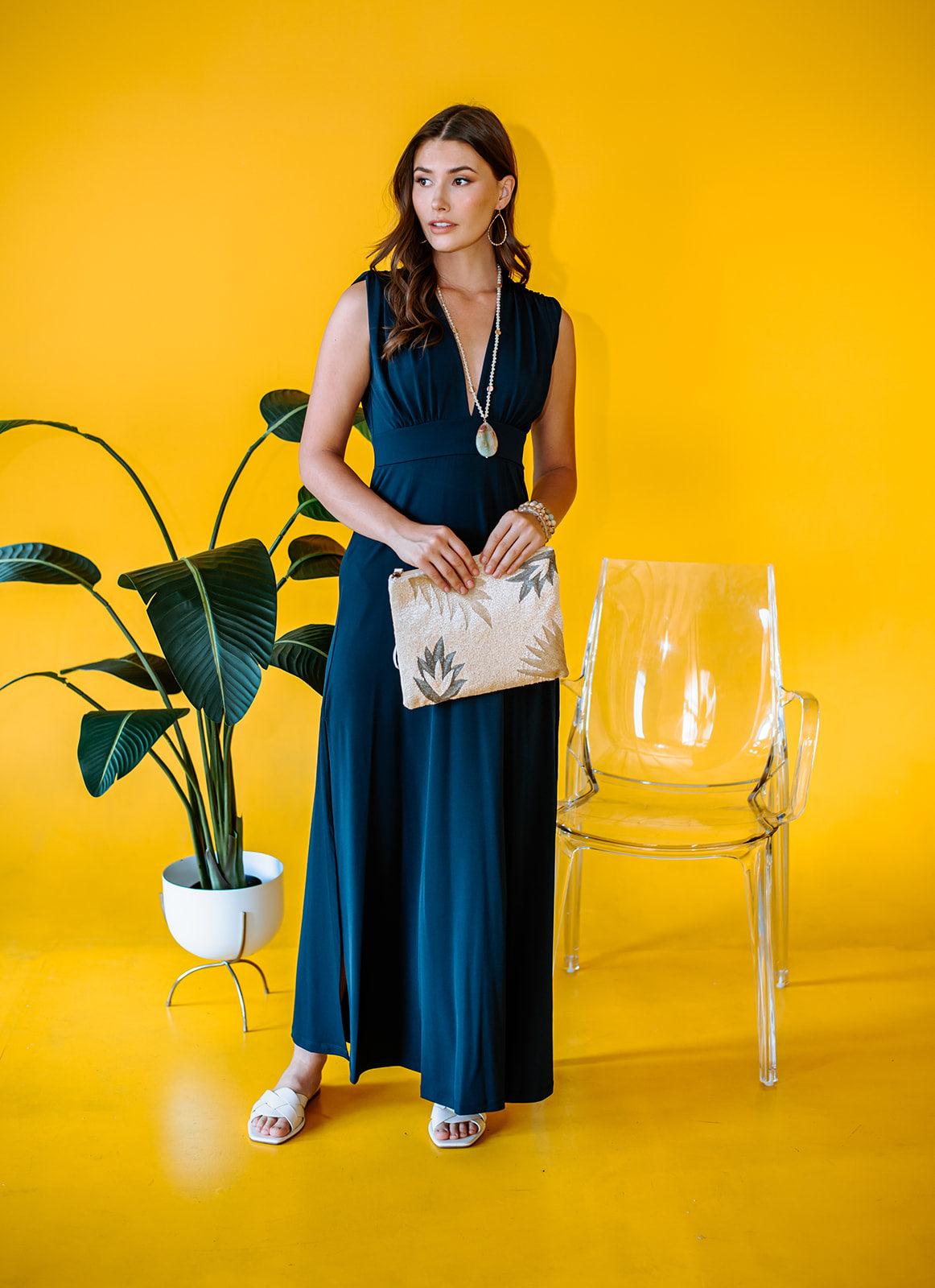 navy sleeveless maxi dress with deep neckline and side slit on woman holding a purse with yellow background