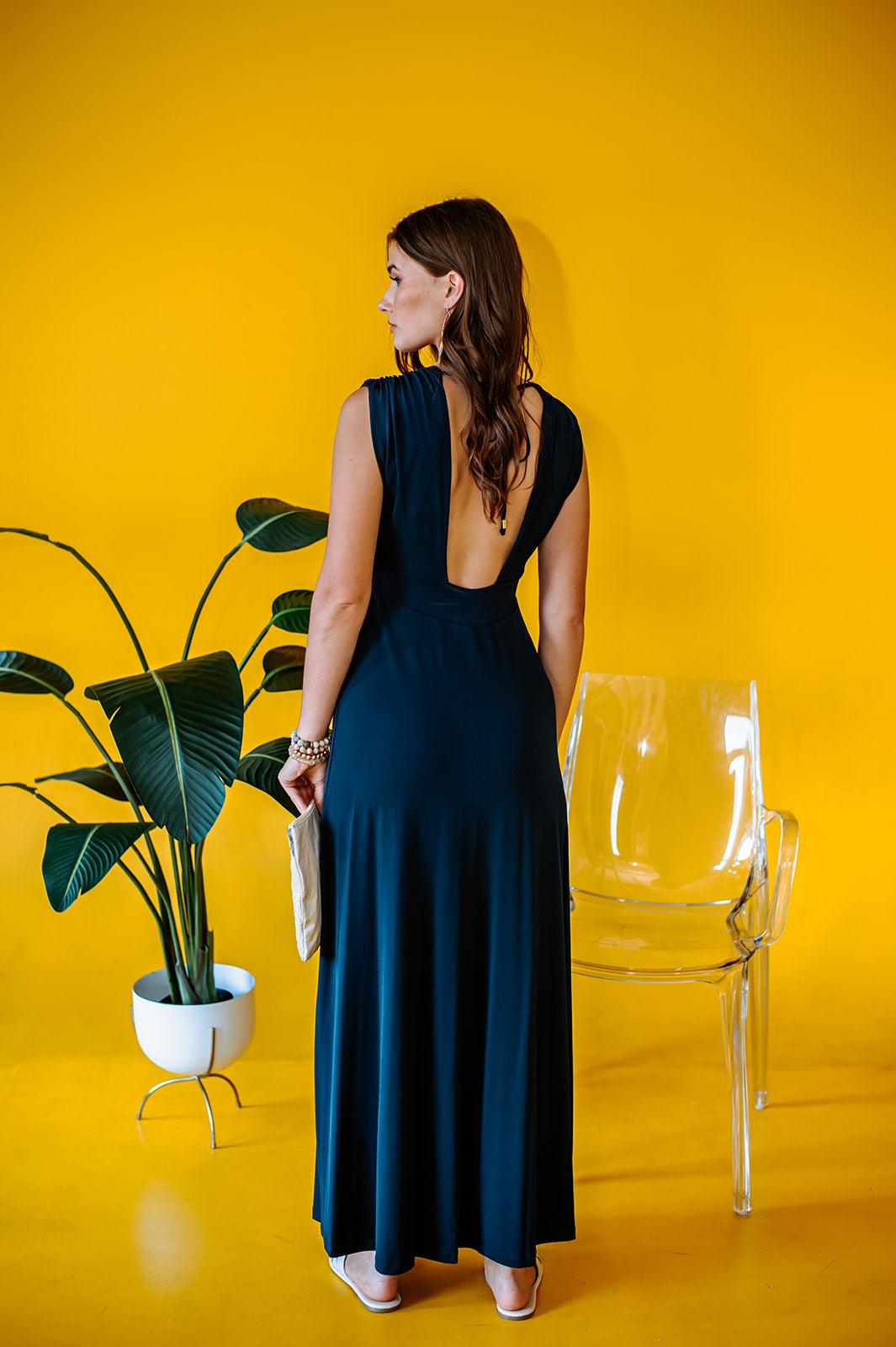 back side of navy sleeveless maxi dress with deep neckline and side slit on woman holding a purse with yellow background