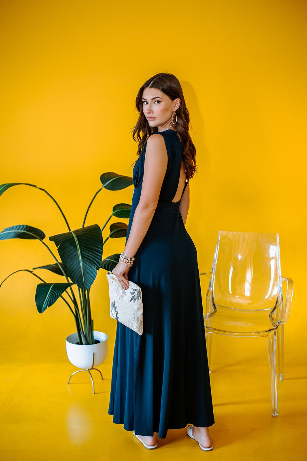 backside of navy sleeveless maxi dress with deep neckline and side slit on woman with yellow background