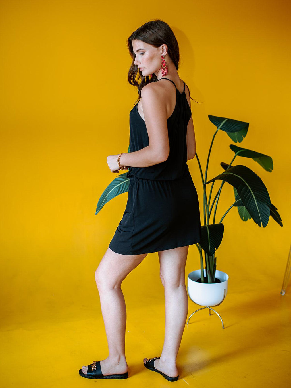 back side of black sleeveless halter dress on woman in black sandals with yellow background